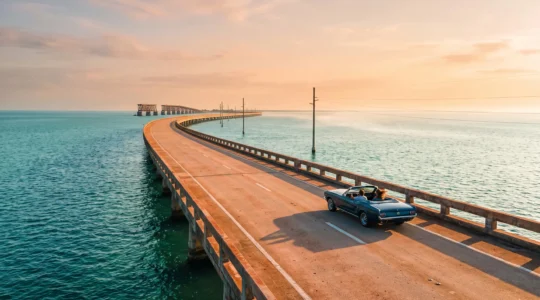 A classic convertible car driving alone on a narrow bridge highway surrounded by turquoise ocean on the way to Key West at golden hour