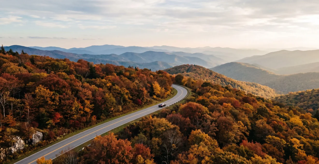 A winding mountain parkway lined with vibrant red, orange and gold trees under a soft morning sky.
