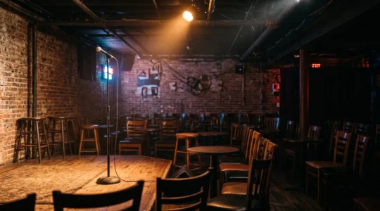 A dimly lit underground comedy club stage with a single microphone under a warm spotlight in an empty brick-walled room before showtime
